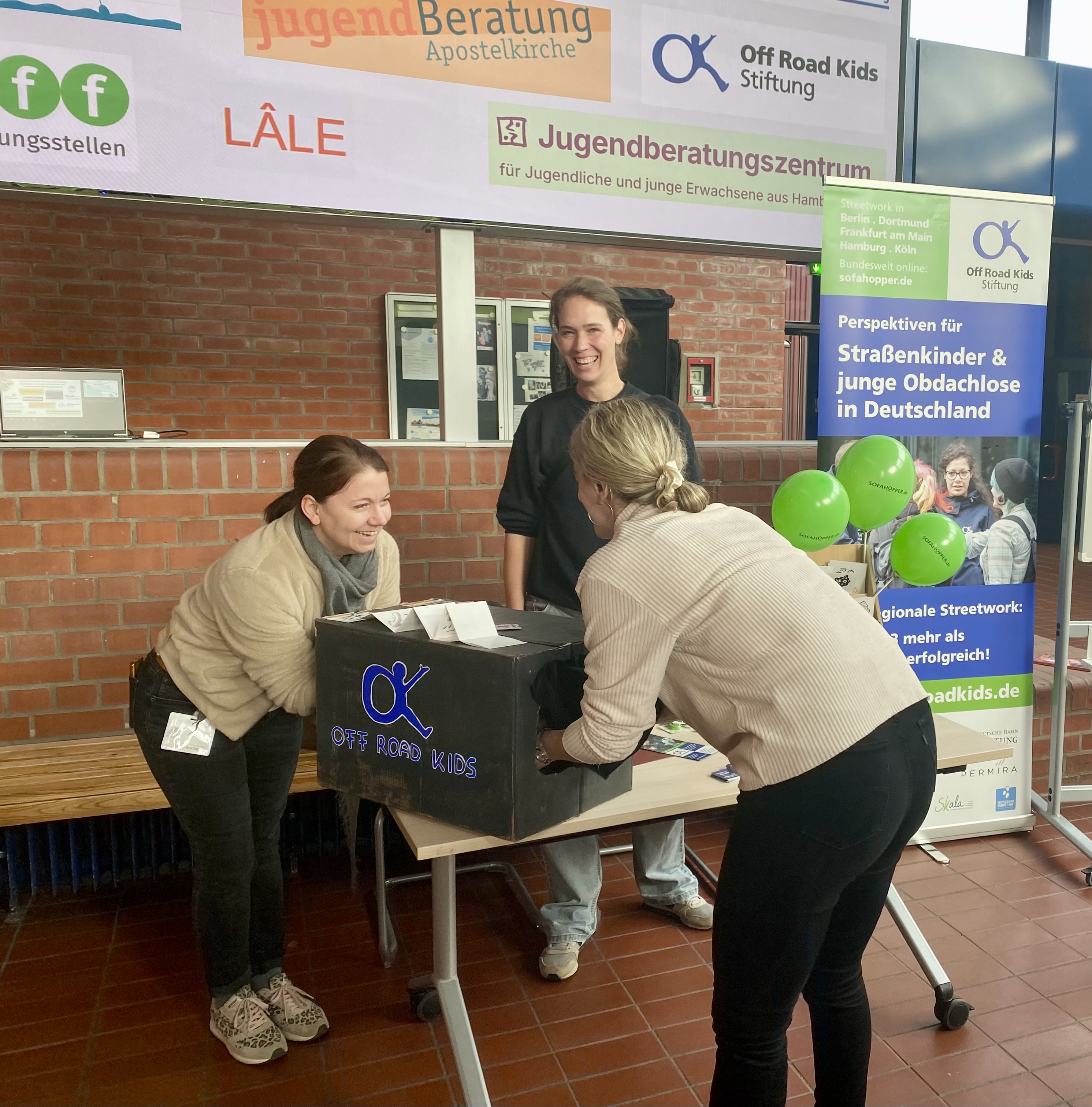 Zwei Personen stehen an einem Informationsstand und fühlen zusammen mit ihren Händen in einer Box, ohne hineinsehen zu können. 