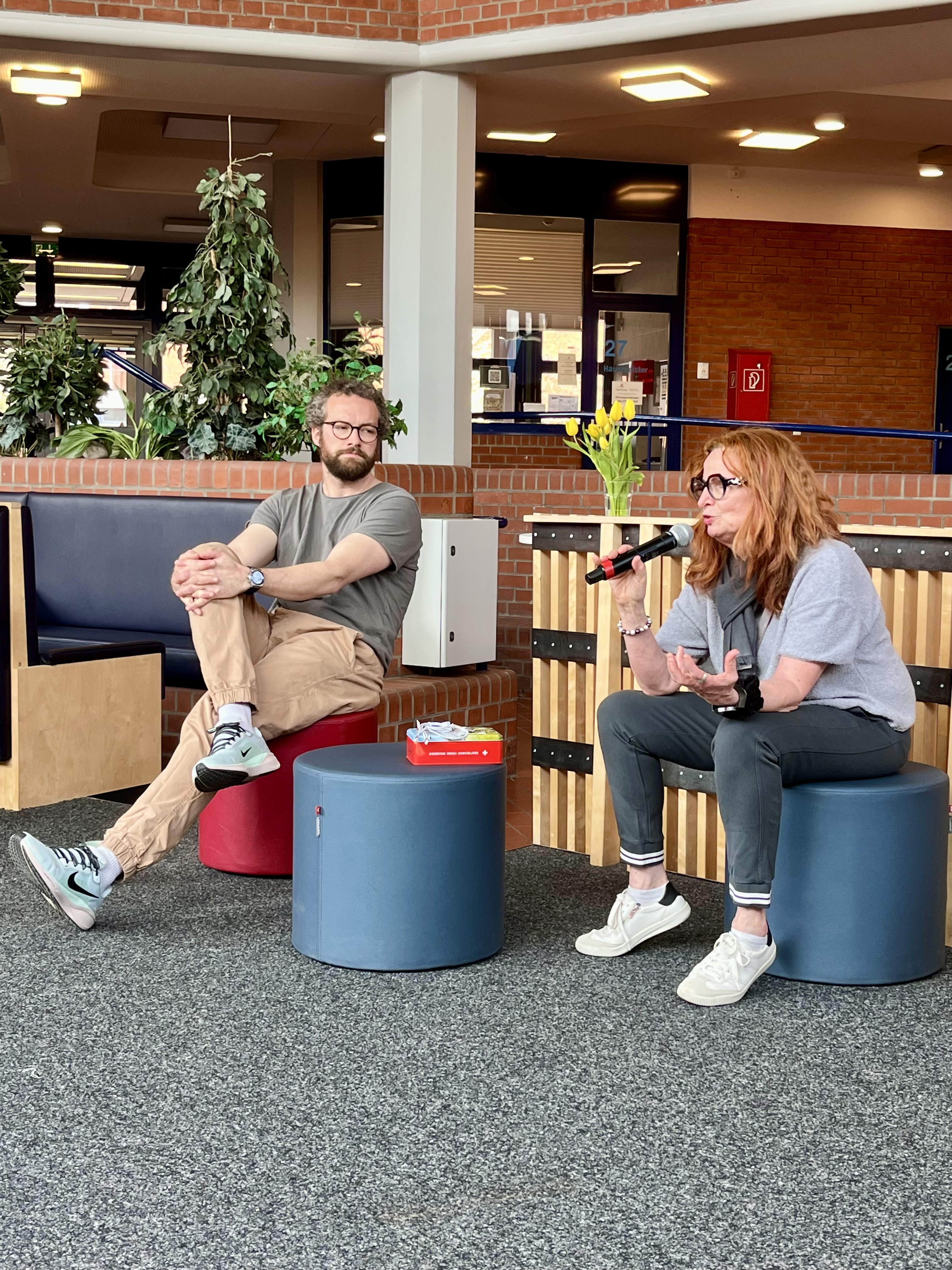 Monika Stausberg sitzt im Atrium uns spricht in ein Mikrofon. Holger Schmidt sitzt daneben. 
