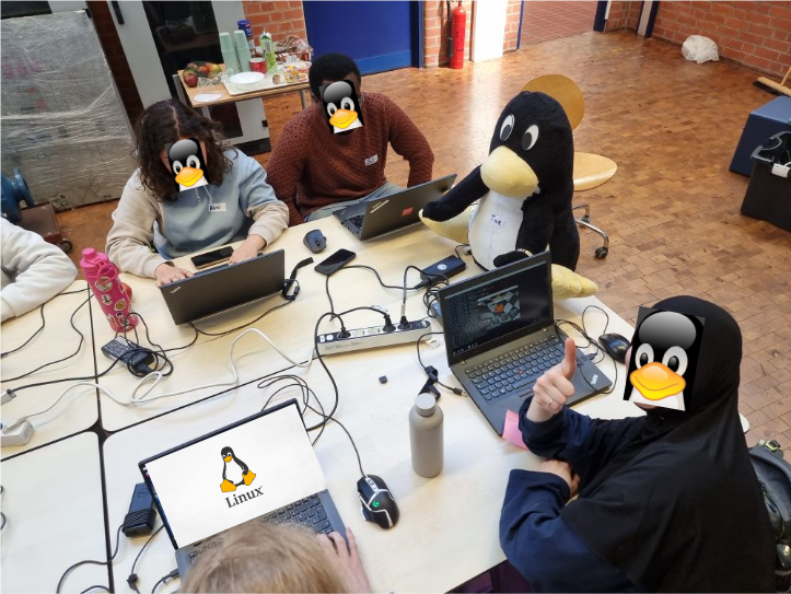 Schüler und Schülerinnen sitzen an einem tisch mit einem Linux-Pinguin-Kuscheltier und arbeiten mit Linusx an Laptops